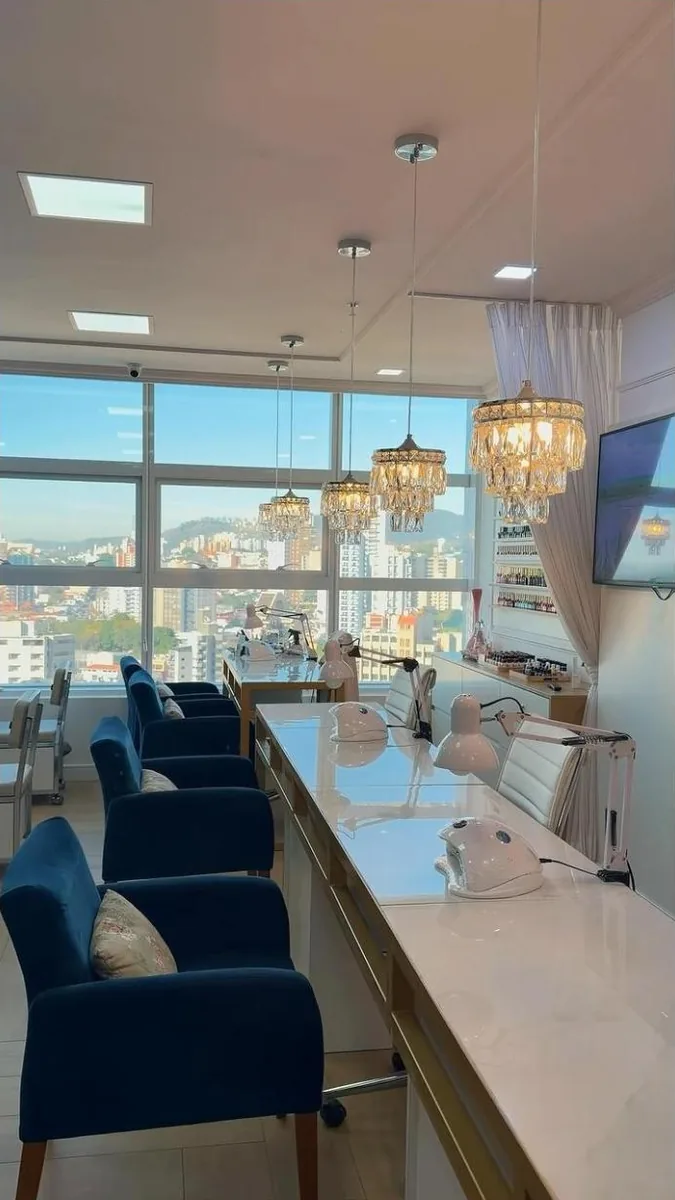 Salão da esmalteria com poltronas azuis, lustres e janelas para a cidade